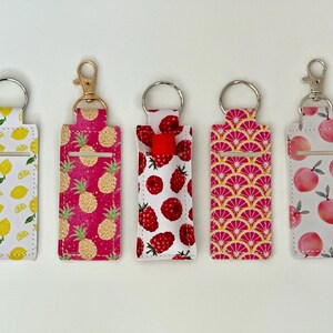 May include: Five fabric lip balm holders with keychains. The patterns include lemons, pineapples, raspberries, pink citrus slices, and peaches. Each holder has a silver or gold-colored keychain ring and clasp.