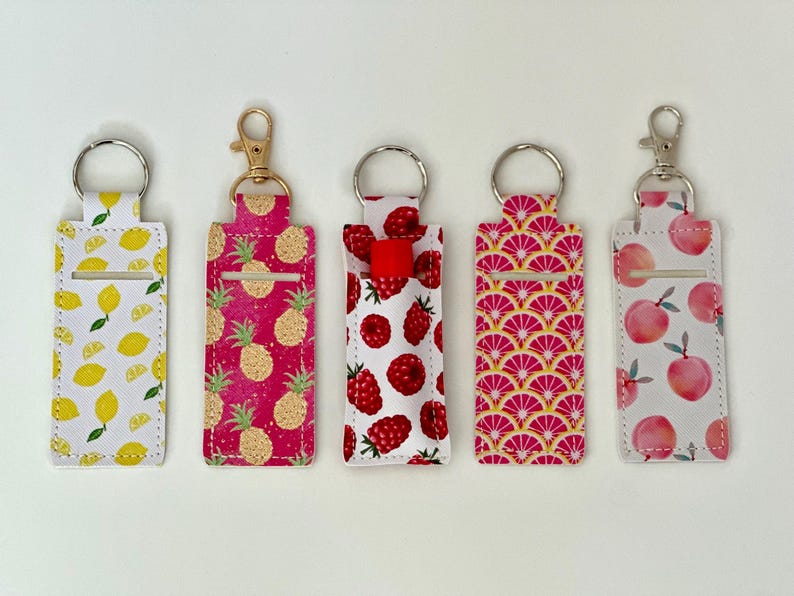 May include: Five fabric lip balm holders with keychains. The patterns include lemons, pineapples, raspberries, pink citrus slices, and peaches. Each holder has a silver or gold-colored keychain ring and clasp.
