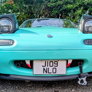 May include: A teal-colored sports car with pop-up headlights. The car has a license plate that reads "JI09 NLO". The front bumper has a unique design with red and white accents. The car is parked outdoors.