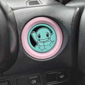 以下が含まれることがあります： ピンクとターコイズの円形フレームの中に、ターコイズ色のゼニガメのデザインが施されたカスタムカーのダッシュボードボタン。ボタンは黒いダッシュボードにセットされており、下には他のボタンが見えます。