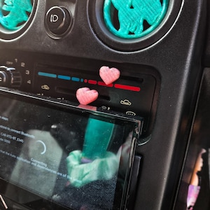 Könnte beinhalten: Ein Armaturenbrett mit tealfarbenen Luftauslassdekorationen in Form eines Pferdes und eines Baumes. Zwei kleine herzförmige Dekorationen in Rosa sind auf dem Armaturenbrett platziert. Der Bildschirm des Autoradios zeigt das Wort "Connecting".