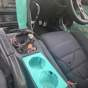 Pode incluir: Acessórios interiores de carro, incluindo um manípulo de mudança de velocidades e um porta-copos turquesa. A bota da alavanca de mudanças é preta com um design floral. O porta-copos tem duas aberturas circulares. O volante é preto.