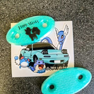 Puede incluir: Dos accesorios ovalados de color turquesa con dos agujeros cada uno, y una pegatina con un coche azul claro y personajes de dibujos animados. Los accesorios tienen el texto "J109 Misty" y una silueta negra.