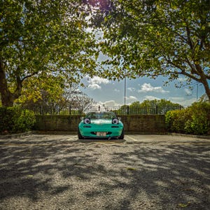 May include: A teal convertible car with cartoon-like eyes on the headlights is parked in a parking lot. The car is positioned in front of a brick wall and under a canopy of green trees. The license plate is visible.