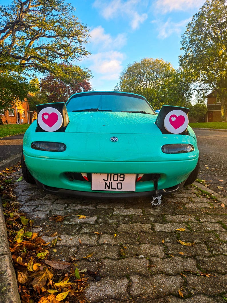 May include: A teal-colored sports car with heart-shaped headlights and a license plate that reads "JI09 NLO." The car has a custom front end with a tooth-like design. The background features trees and a blue sky.