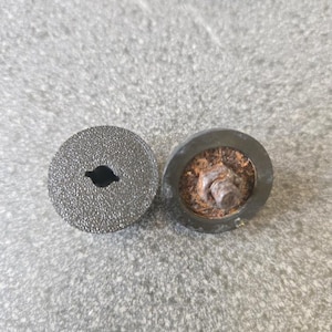 May include: Two small, dark grey, circular objects with a textured surface. One has a keyhole-shaped opening in the centre. The other has a rusty bolt and washer attached. The objects are on a light grey surface.
