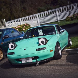 Puede incluir: Un coche deportivo turquesa con ojos de dibujos animados en los faros. El coche tiene un techo negro, una matrícula delantera y un gancho de remolque. El coche está aparcado sobre asfalto cerca de una valla blanca.