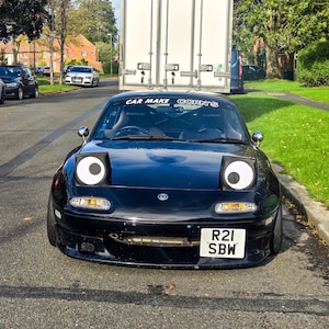 May include: A black sports car with large, cartoon-like eyes on the headlights, parked on a road. The car has a license plate that reads "R21 SBW". Above the windshield is the text "CAR MAKE CORN'S". A white truck is in the background.