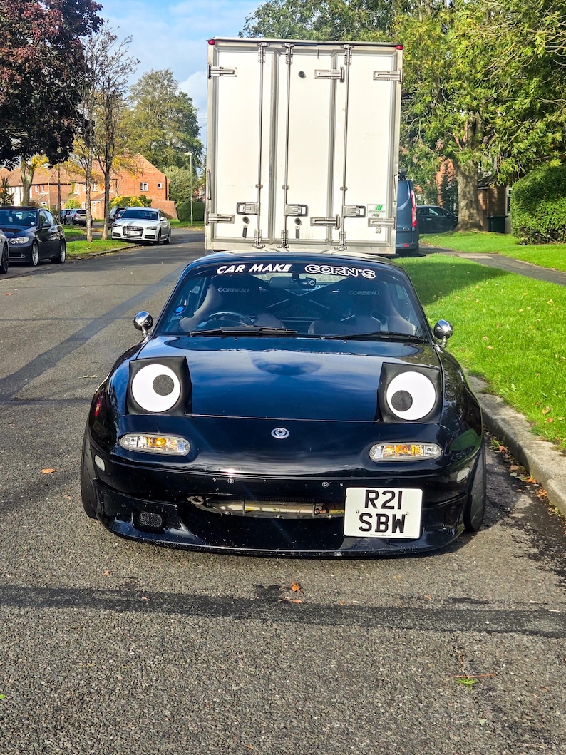 May include: A black sports car with large, cartoon-like eyes on the headlights, parked on a road. The car has a license plate that reads "R21 SBW". Above the windshield is the text "CAR MAKE CORN'S". A white truck is in the background.