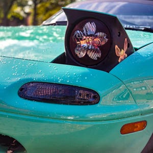 May include: A teal car with a pop-up headlight featuring a cherry blossom petal design. The bonnet is covered in water droplets, and a small cartoon character sticker is visible on the headlight housing. A dark turn signal and orange side marker are present.