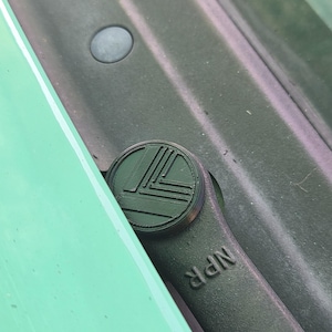 May include: A dark green windscreen wiper arm featuring a circular cap with a geometric pattern of raised lines. The arm has the letters NPR embossed on its surface. The component is positioned against a light green vehicle body panel.