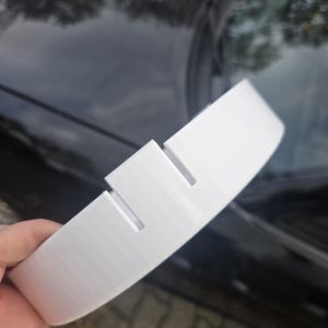 May include: A white, curved plastic object with two rectangular cutouts. The object appears to be a car part, possibly a spoiler or trim piece. The item is held against a dark, reflective surface.