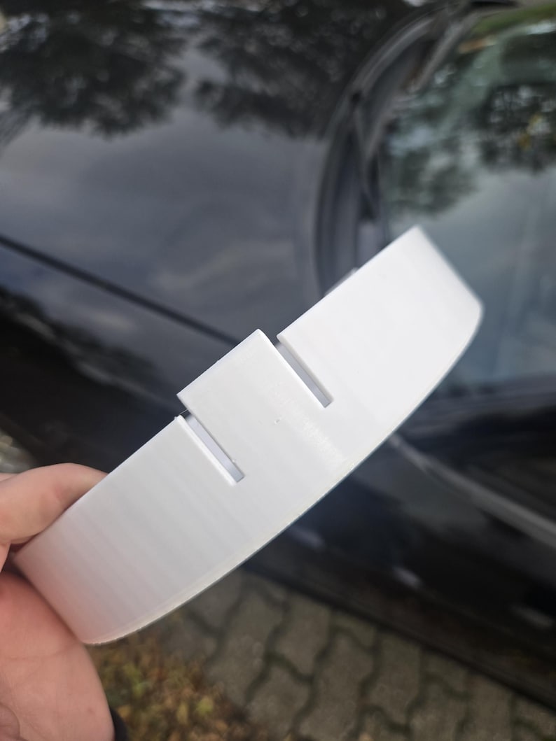 May include: A white, curved plastic object with two rectangular cutouts. The object appears to be a car part, possibly a spoiler or trim piece. The item is held against a dark, reflective surface.