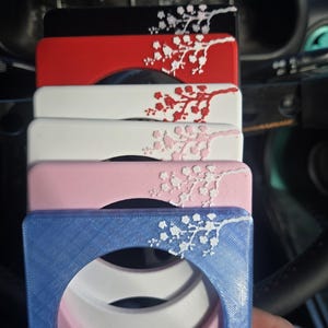 May include: A selection of car coasters in black, red, white, pink, and blue. Each coaster has a square frame with a circular centre and a white floral design. The coasters are in a car.