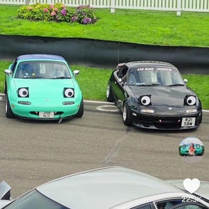 May include: Two Mazda Miata cars, one teal and one black, with cartoon-like eyes on the headlights. The teal car has a license plate that reads "JI09 NLO". The black car has a license plate that reads "R21 SBW".
