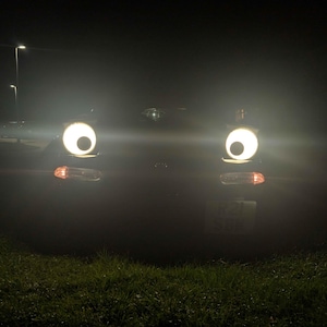 May include: A car's headlights at night, appearing as large, bright eyes. The headlights are round and glowing, with a dark background. The car's turn signals are also visible. The scene is set on a grassy area with streetlights in the distance.