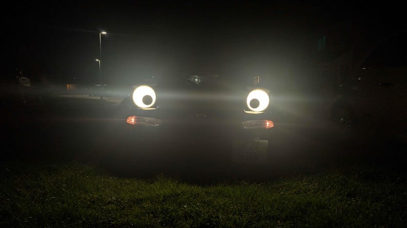 May include: A car's headlights at night, appearing as large, bright eyes. The headlights are round and glowing, with a dark background. The car's turn signals are also visible. The scene is set on a grassy area with streetlights in the distance.