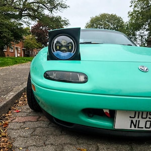 May include: A teal sports car with a pop-up headlight. The headlight is black and round, with a detailed interior. The car is parked on a brick-paved street, with trees and houses in the background.