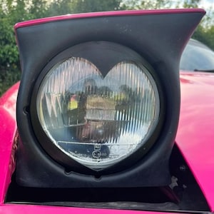 May include: A circular car headlight featuring a black housing with a heart-shaped cutout over the lens. The headlight is mounted on a bright pink vehicle body, with the clear glass lens showing horizontal fluting and small regulatory markings.
