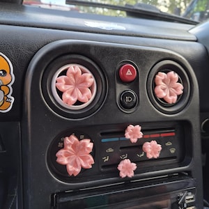 May include: A set of six pink cherry blossom-shaped car air vent clips attached to the dashboard of a vehicle. Two larger clips are positioned inside the circular air vents, while four smaller clips are secured to the horizontal vent slats.