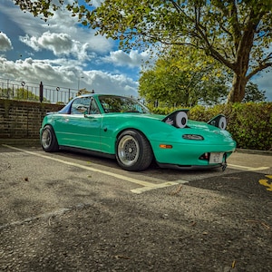 May include: A teal convertible car with black rims is parked in a parking lot. The car has large, cartoonish googly eyes attached to the front. The sky is blue with fluffy white clouds, and a tree is in the background.