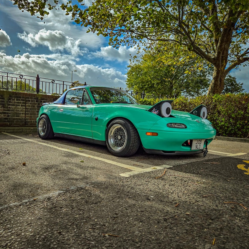 May include: A teal convertible car with black rims is parked in a parking lot. The car has large, cartoonish googly eyes attached to the front. The sky is blue with fluffy white clouds, and a tree is in the background.