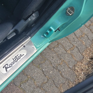Peut inclure: Intérieur d'une portière de voiture turquoise avec un siège noir visible. La portière présente une plaque argentée avec le mot "Roadster" en écriture cursive. La portière est ouverte et possède une poignée noire.