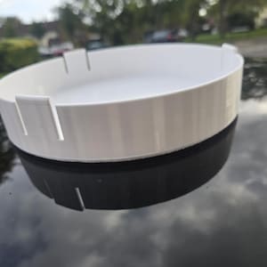 May include: A white, circular tray with a raised rim and evenly spaced rectangular cutouts. The tray sits atop a black, reflective surface. The design is simple and modern, suitable for various uses.