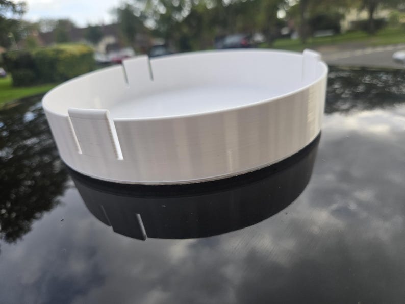 May include: A white, circular tray with a raised rim and evenly spaced rectangular cutouts. The tray sits atop a black, reflective surface. The design is simple and modern, suitable for various uses.