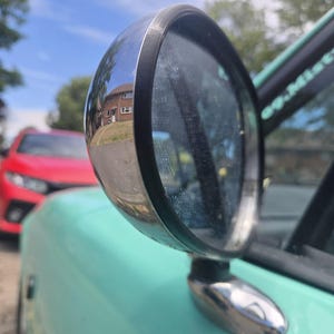 Puede incluir: Espejo retrovisor cromado con un borde de goma negro, montado en un coche verde azulado claro. El espejo refleja una escena residencial con un coche rojo en el fondo. El cielo es azul con nubes.