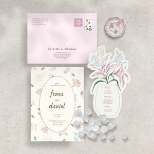 Peut inclure: Ensemble de papeterie de mariage comprenant une enveloppe rose avec des timbres floraux, une invitation florale et une carte en forme de vase avec un motif floral. Des p&eacute;tales de fleurs blancs sont dispers&eacute;s.