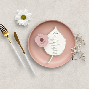 Peut inclure: Une composition &agrave; plat avec une assiette rose, des couverts dor&eacute;s et blancs, et une marguerite blanche. Un menu en forme de feuille et une carte rose en forme de fleur avec du texte sont sur l'assiette. Le menu indique "Let's eat".