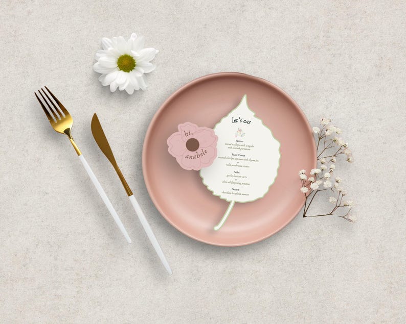Peut inclure: Une composition &agrave; plat avec une assiette rose, des couverts dor&eacute;s et blancs, et une marguerite blanche. Un menu en forme de feuille et une carte rose en forme de fleur avec du texte sont sur l'assiette. Le menu indique "Let's eat".