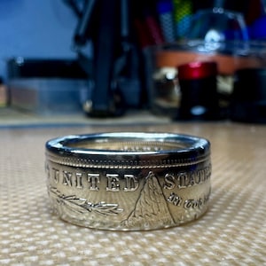 May include: A silver ring made from a United States half dollar coin. The ring features the words "United States of America" and "In God We Trust" on the side. The ring is a unique and stylish accessory.