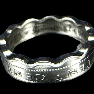 May include: A silver ring with a wavy edge and engraved text. The ring appears to be made from a coin, with visible details of the original coin's design. The ring is set against a black background.