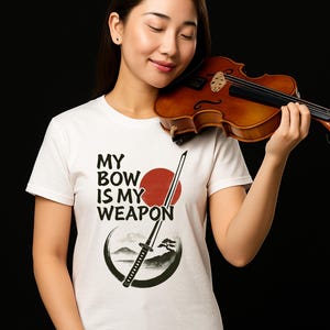 Puede incluir: Camiseta blanca con el texto "MY BOW IS MY WEAPON" en negro, con un sol rojo y un gráfico de espada. Una persona sostiene un violín marrón. El fondo es negro.