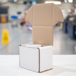 K&ouml;nnte beinhalten: Eine wei&szlig;e Pappbox mit einem braunen Pappdeckel. Der Deckel ist ge&ouml;ffnet und die Box ist leer.