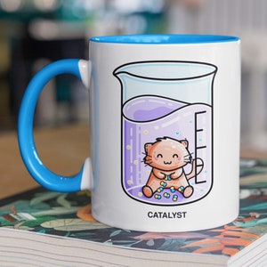May include: White ceramic mug with a blue rim and handle. The mug features a cartoon illustration of a cat inside a beaker with purple liquid and the word "CATALYST" printed below.