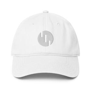 May include: White baseball cap with a curved peak and adjustable strap. The cap has a grey circular embroidered logo on the front. The logo is a stylized design with vertical and horizontal lines.