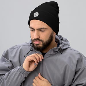 May include: A black beanie hat with a white circular logo. The beanie has a ribbed texture and is made of a soft material. The logo is a stylized letter "S".
