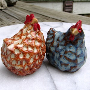 May include: Two ceramic chicken figurines. One is brown and white with a red comb and wattle. The other is blue and brown with a red comb and wattle.