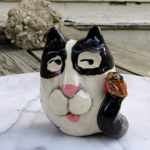 May include: A ceramic figurine of a black and white cat with a pink nose and a small brown bird perched on its arm.