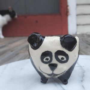 May include: A ceramic panda bear planter with a black and white design. The planter has a smiling face and small legs. The ears and details are black, while the face is white. A cat is visible in the background.