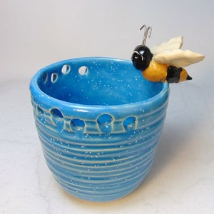 May include: A blue ceramic planter with a bee figurine on the rim. The planter has a speckled glaze and a ribbed design. The bee is yellow and black with white wings.