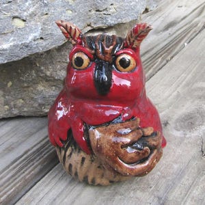 May include: A ceramic owl figurine with a glossy red body, brown wings, and a black beak. The owl has large, yellow eyes and small, pointed ears. The figurine is sitting on a wooden surface.