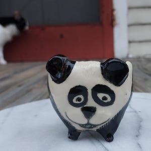 May include: A small ceramic panda figurine with black and white paint. The panda has a cute expression with big eyes and a small nose.