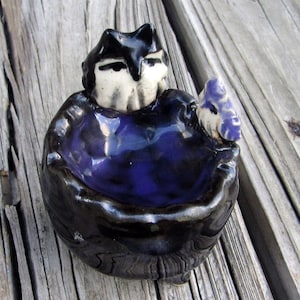 May include: A small ceramic dish with a black and purple glaze. The dish features a black cat figurine with white eyes and a purple flower.