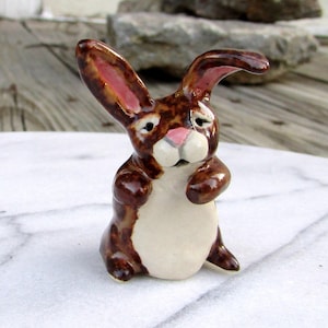May include: A ceramic bunny figurine with brown and white coloring. The bunny has long ears with pink inner ears, a pink nose, and is sitting upright. The figurine is on a white marble surface.