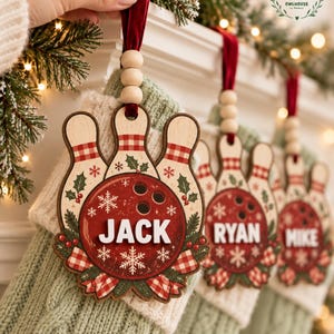 May include: A wooden bowling-themed Christmas stocking tag featuring a red bowling ball with three finger holes, white snowflakes, and holly accents. The name JACK is printed in white block letters. It hangs from a red velvet ribbon with three wooden beads.