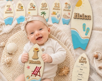 May include: A set of wooden surfboard-shaped milestone markers featuring numbers 1 through 12, a personalised name plaque with the name Helen, and a birth announcement plaque with fields for date, time, weight of 7lbs 4oz, and length of 20.5 inches.